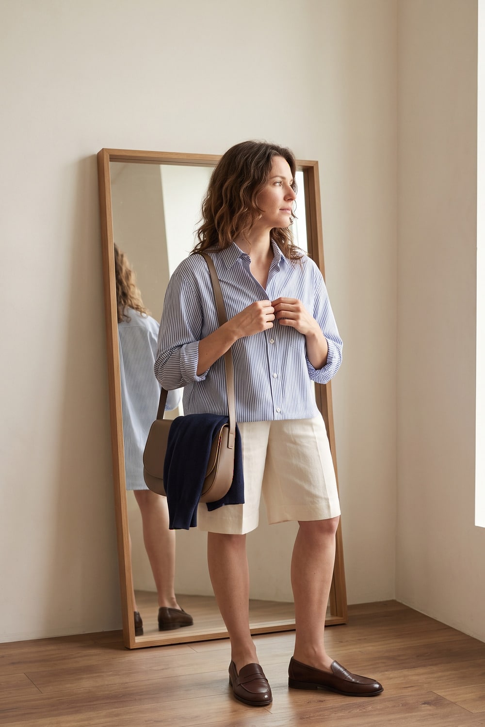 Soft Preppy Style for Late Summer Woman in a soft preppy style outfit with a pale blue shirt tailored long shorts loafers and a light cardigan for late summer